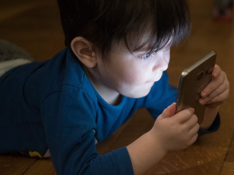 A young boy lying on a wooden floor looks closely at a smartphone screen