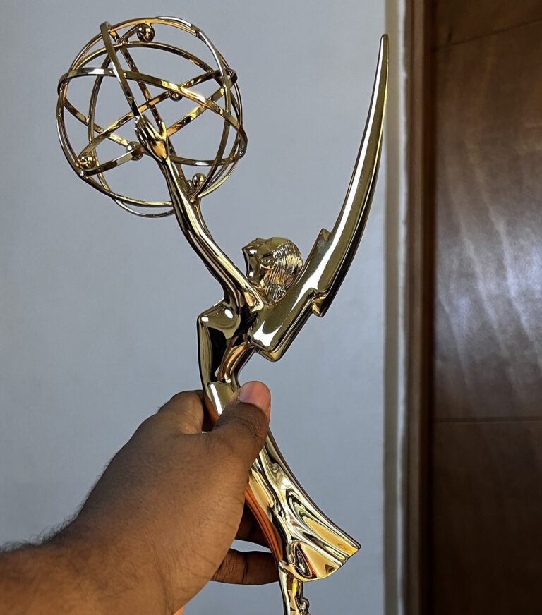 A hand holding a gold Emmy Award statuette against a plain wall