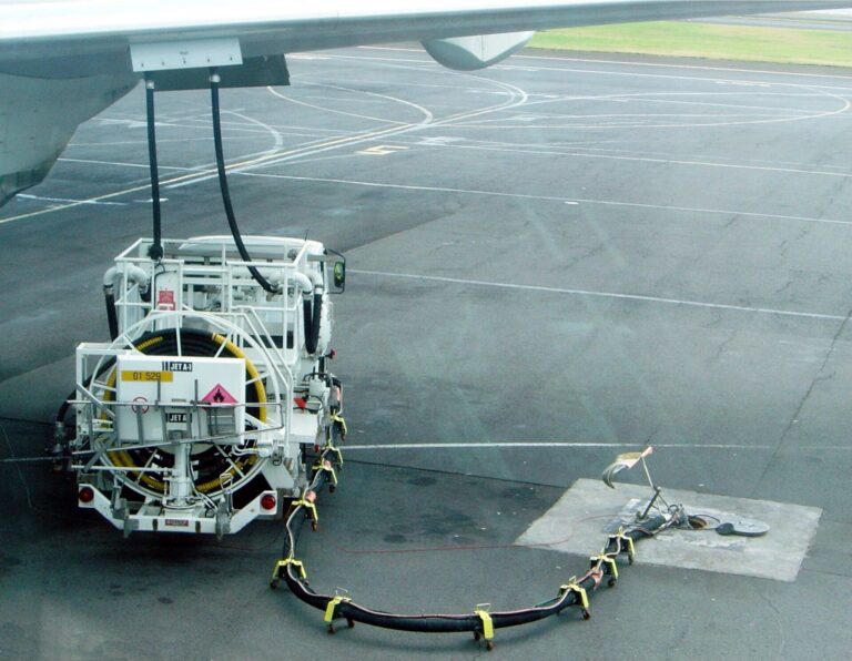 A fuel service truck connected by hoses to the wing of a parked airliner on an airport apron